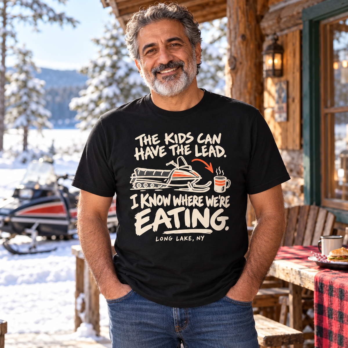 Older rider wearing The Kids Can Have the Lead I Know Where We're Eating Long Lake snowmobile tee at a snowy cabin lunch stop.