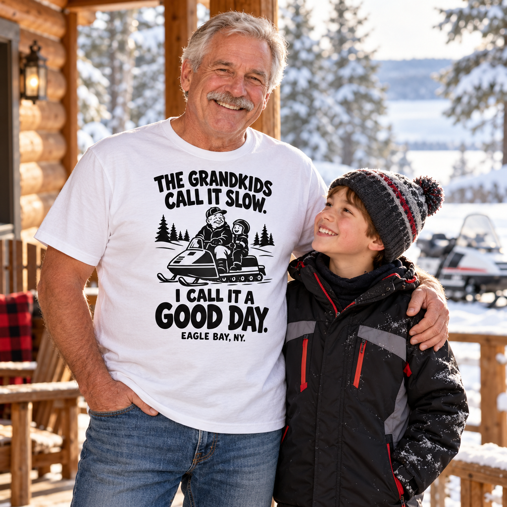 Grandfather wearing Eagle Bay Good Day snowmobile tee with grandchild on a snowy cabin porch.