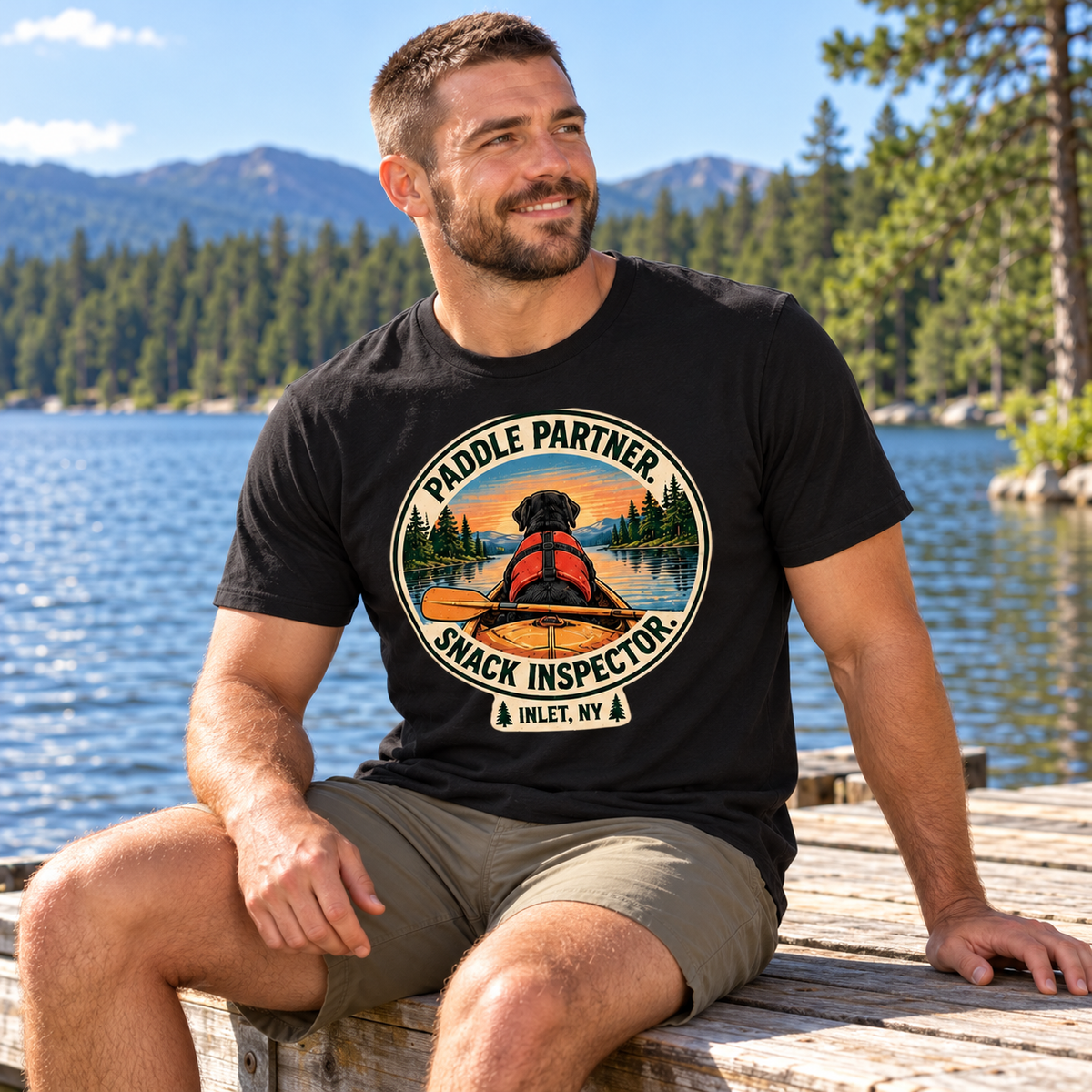 Bearded man seated on a dock wearing Paddle Partner Snack Inspector tee
